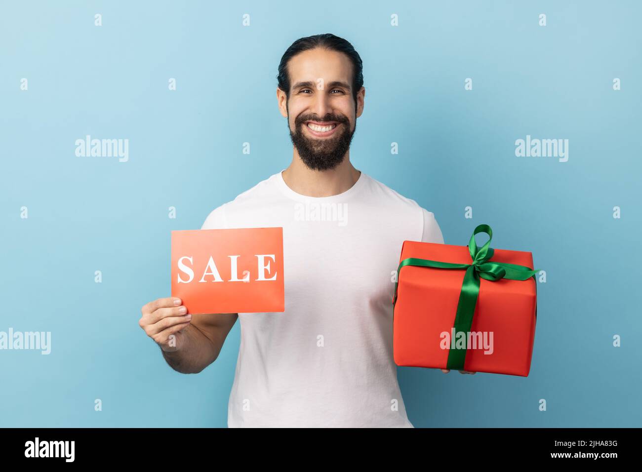 Bonus sul Black Friday shopping. Ritratto dell'uomo che indossa una T-shirt bianca che mostra l'iscrizione alla vendita e la confezione regalo, guardando la macchina fotografica con un sorriso toothy. Studio interno girato isolato su sfondo blu. Foto Stock