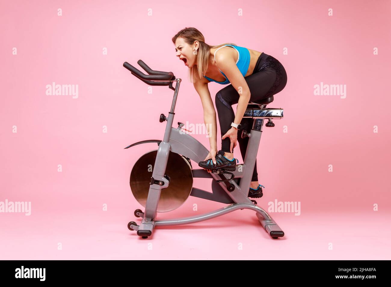 Il ritratto completo di sportswoman si è allenato a casa sulla bici da sforzo senza scaldarsi e ferito la sua gamba, indossando i tights sportivi e la parte superiore. Studio interno girato isolato su sfondo rosa. Foto Stock