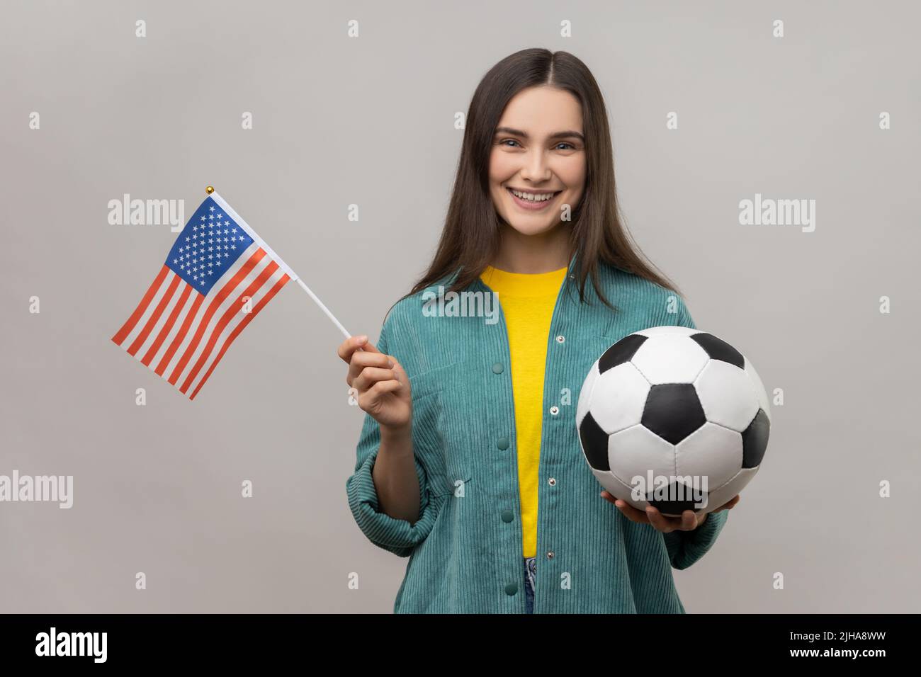 Donna sorridente che tiene bandiera degli stati uniti d'america e calcio palla bianca e nera, lega di calcio degli stati uniti, indossando giacca casual stile. Studio interno girato isolato su sfondo grigio. Foto Stock