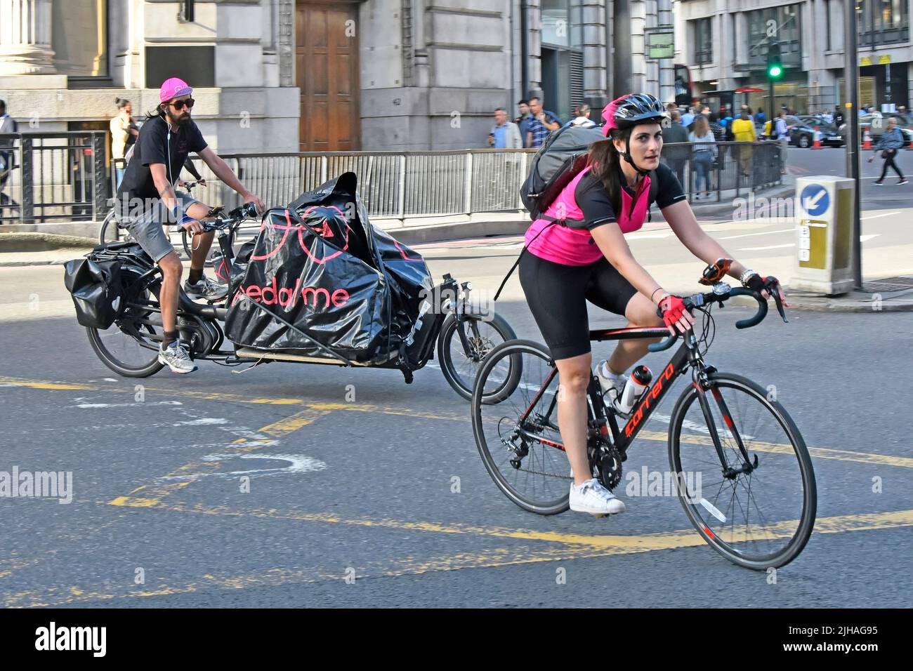 Donna ciclista con zaino che guida drop handle bar bicicletta attraverso City of London box Junction Road seguito da un uomo sulla lunga bicicletta da carico Inghilterra UK Foto Stock
