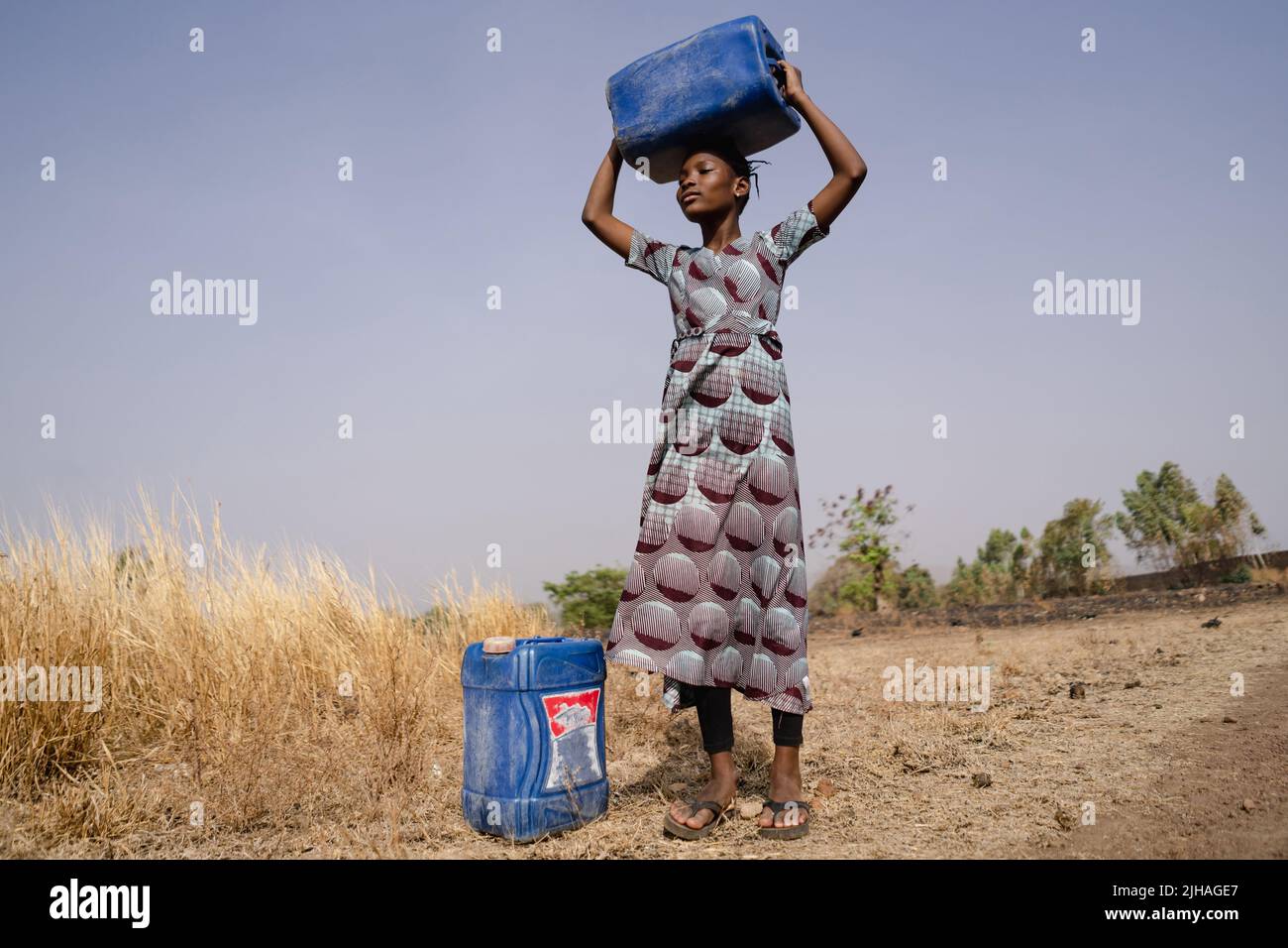 Ragazza africana skinny con i contenitori di acqua che guardano in lontananza, stimando la lunga passeggiata al rubinetto del villaggio Foto Stock
