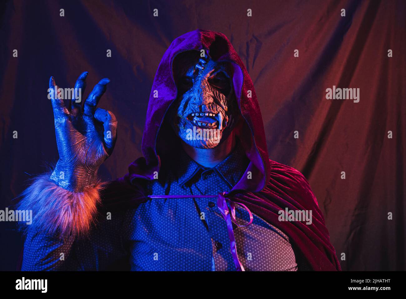 Ritratto di uno zombie vestito in una camicia e mantello con cappuccio facendo un giusto gesto con la mano. La scena è scura, illuminata da luci blu e arancioni Foto Stock