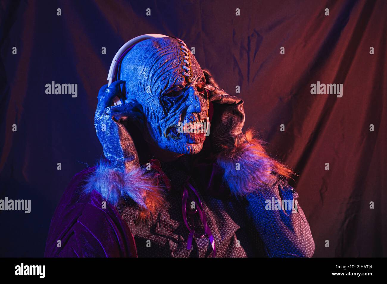 Ritratto di uno zombie vestito con una camicia e mantello ascoltando musica con cuffie wireless. La scena è scura, illuminata da luci blu e arancioni Foto Stock
