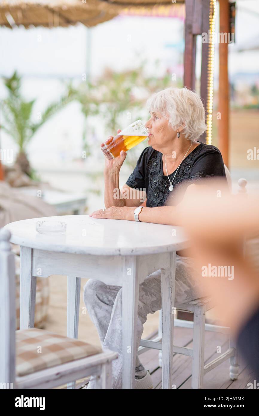 Donna matura seduta a un tavolo in un bar estivo e bere birra Foto Stock