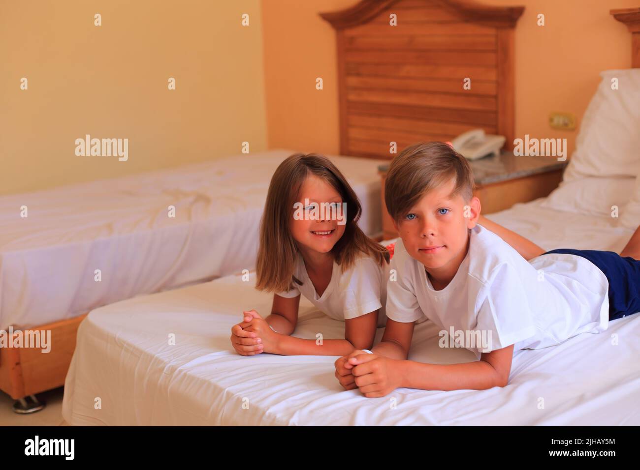 Due bambini si trovano su un letto con lenzuola bianche in una camera d'albergo in vacanza. I bambini giocano sul letto con lenzuola bianche a casa. Ragazzo e ragazza prima di andare a letto a casa Foto Stock