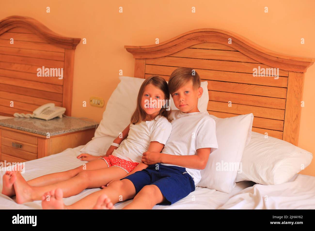 Due bambini si trovano su un letto con lenzuola bianche in una camera d'albergo in vacanza. I bambini giocano sul letto con lenzuola bianche a casa. Ragazzo e ragazza prima di andare a letto a casa Foto Stock