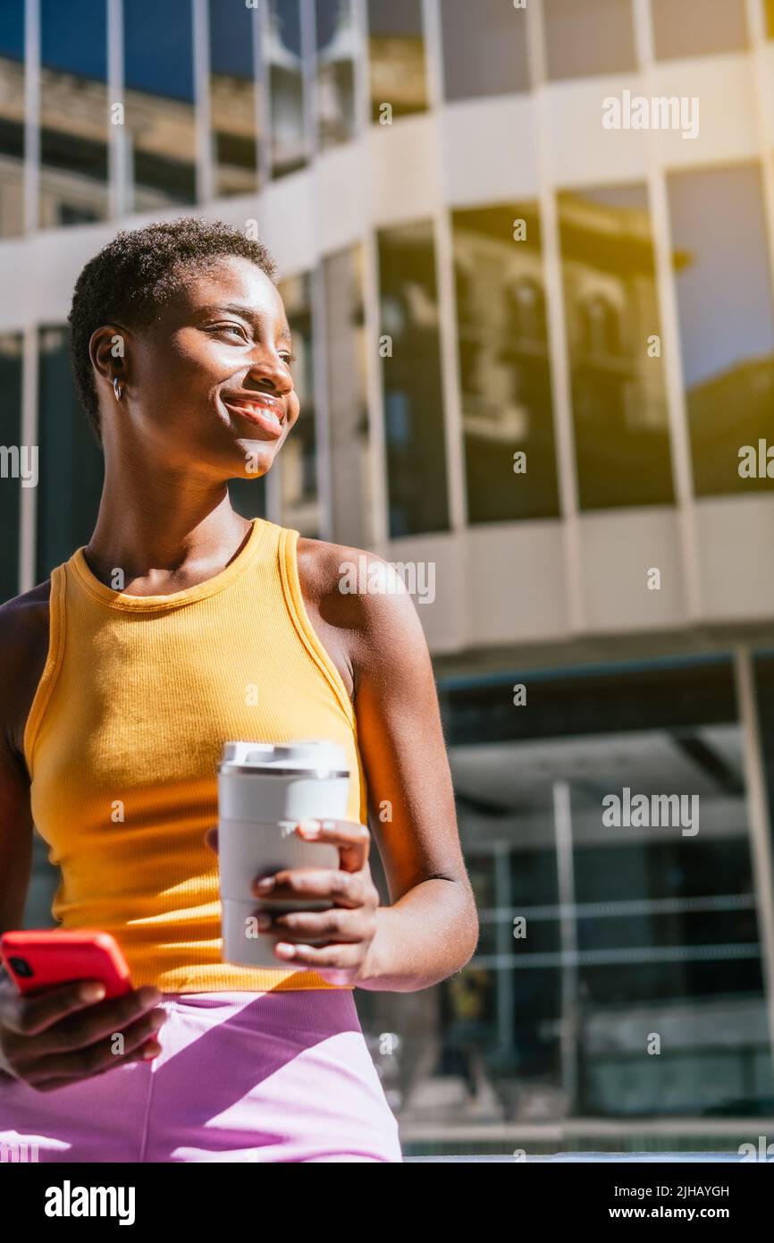 Allegra donna africana con capelli molto corti in attesa in strada. Contiene una tazza da caffè riutilizzabile e uno smartphone. Foto Stock