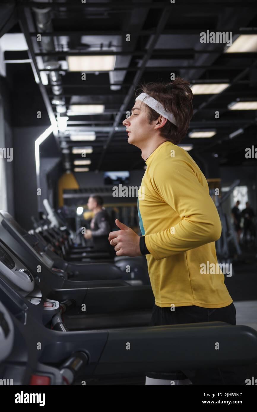 Colpo a profilo verticale di un atleta maschio che indossa la fascia, che corre sul tapis roulant in palestra Foto Stock