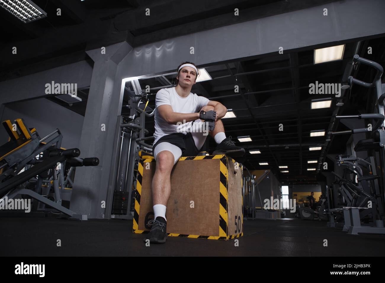 Basso angolo di ripresa a tutta lunghezza di un bel atletico uomo seduto su una scatola di salto in palestra, riposandosi dopo l'esercizio Foto Stock