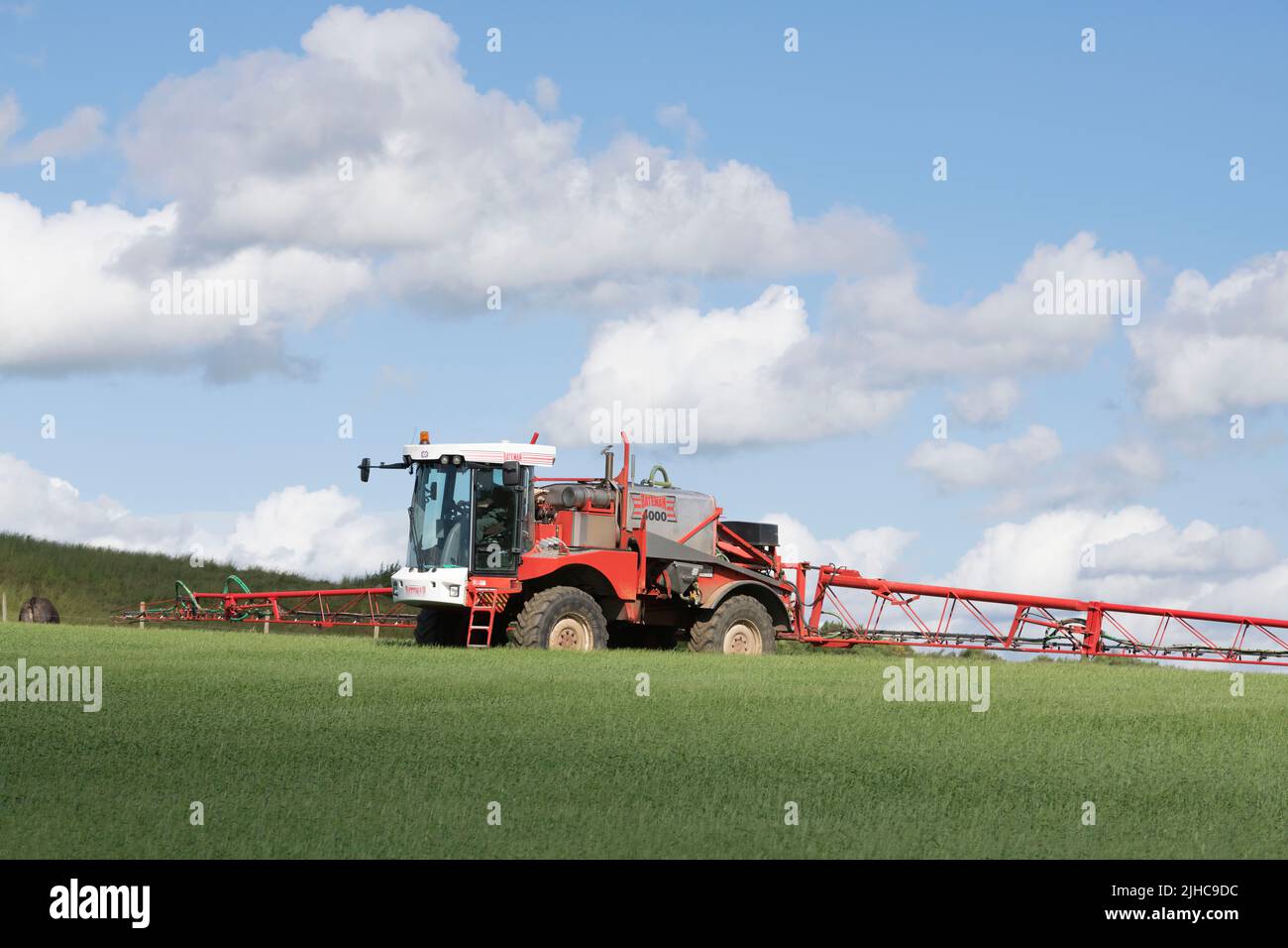 Un irroratore di raccolto che lavora in un campo di Barley di primavera sotto Cumulus Clouds in una mattinata estiva soleggiato Foto Stock