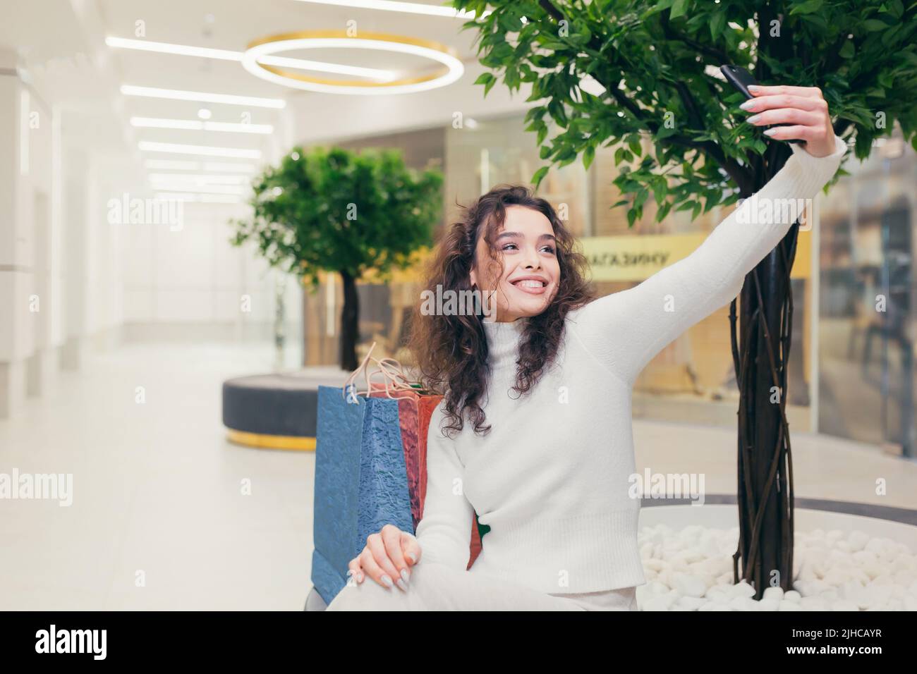 Felice e soddisfatto giovane bella ragazza curly, donna seduta su una panchina nel centro commerciale. Tiene un telefono in mano, prende un selfie. Sorrisi Foto Stock