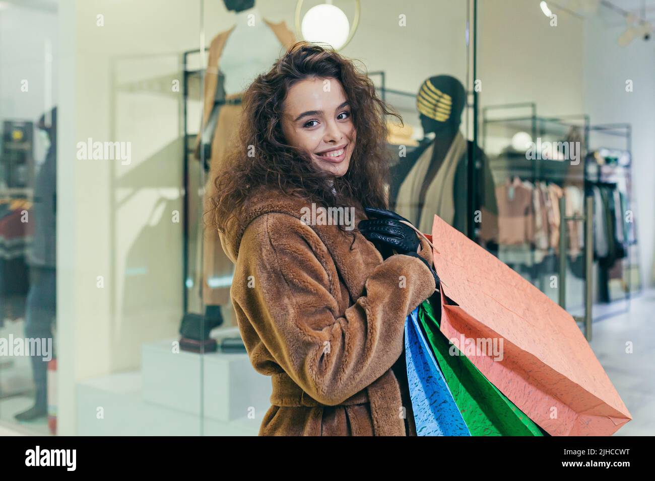 Vendita vacanze. Bella giovane donna ricci in pelliccia cappotto che tiene carta borse colorate con shopping e regali per le vacanze di Natale per la famiglia e gli amici. Spettacoli, brags, gioie, guarda la macchina fotografica in un negozio in un centro commerciale Foto Stock