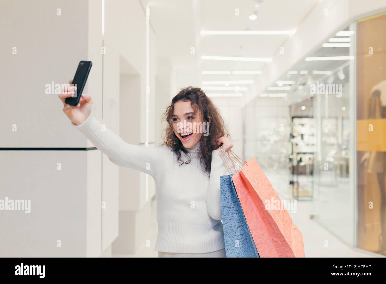 Giovane bella donna shopping in un negozio, prendendo un selfie su un telefono cellulare. Contiene sacchetti colorati con la merce. Ama fare shopping Foto Stock