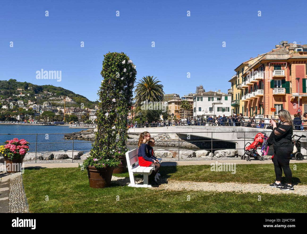 Una donna fotografa ragazze sedute su una panchina tra decorazioni floreali durante le celebrazioni pasquali sul lungomare di Rapallo, Genova, Liguria, Ital Foto Stock