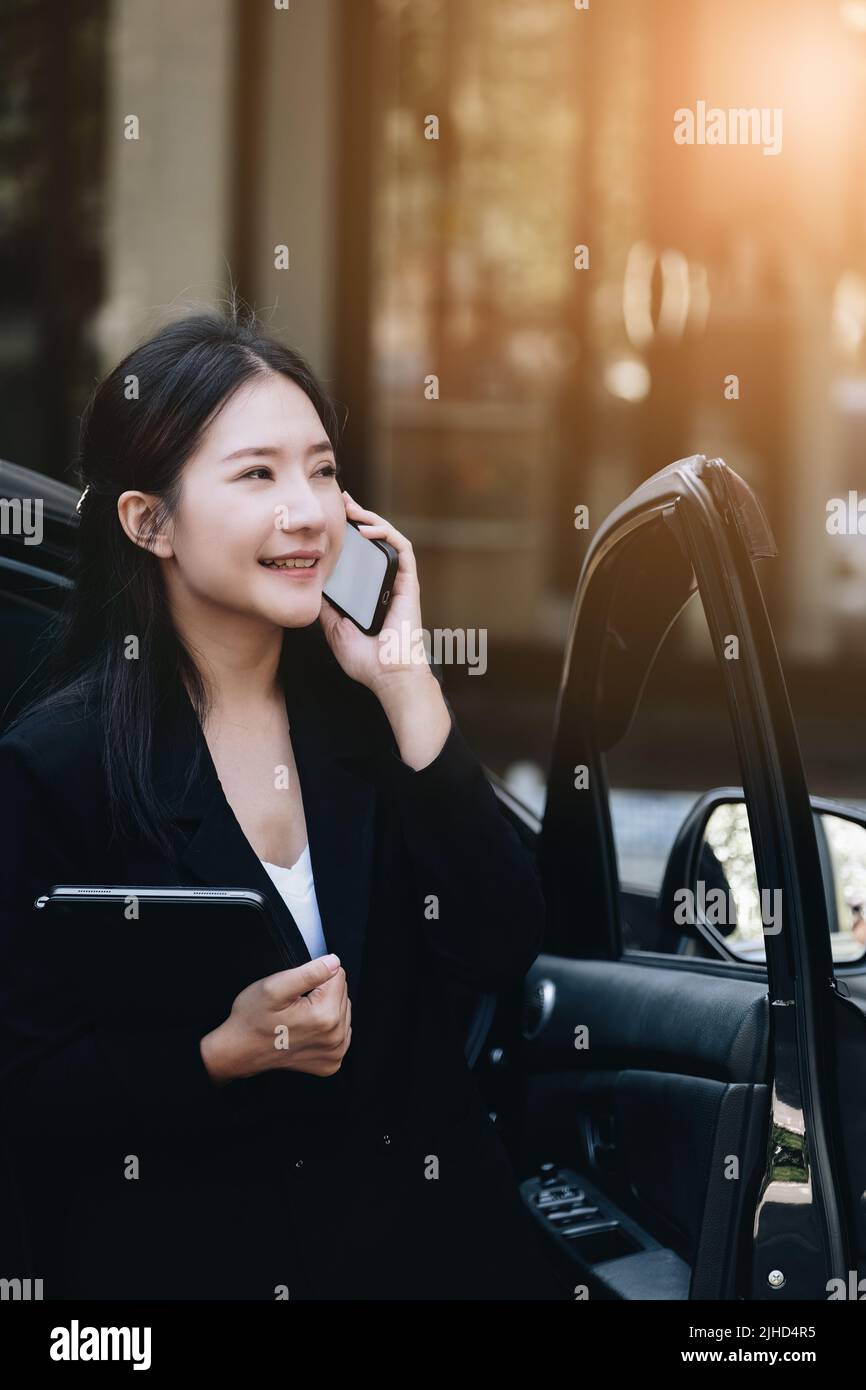 Gli uomini d'affari asiatici, i proprietari di affari, i presidenti di azienda o i dipendenti femminili che parlano al telefono e che tengono un tablet stanno uscendo dall'automobile a. Foto Stock