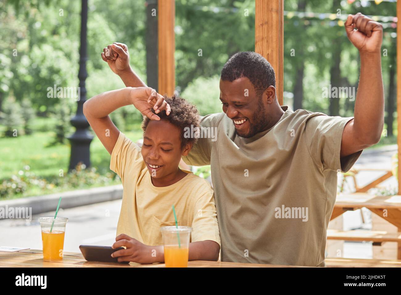 Eccitato papà felice per la vittoria di suo figlio nel gioco online sul telefono cellulare mentre bevono succo al caffè all'aperto Foto Stock