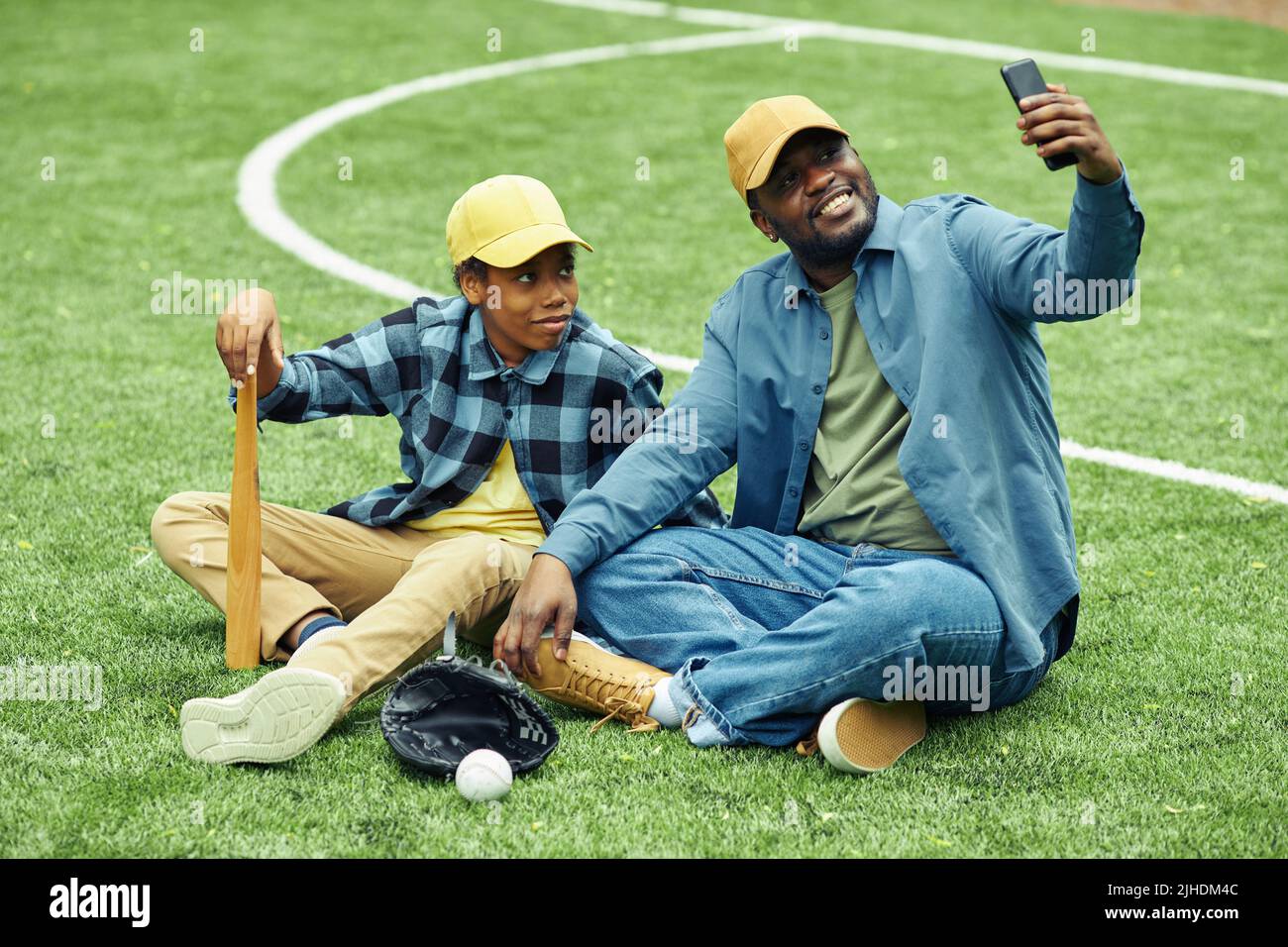 Felice papà che fa ritratto selfie con suo figlio sul suo cellulare mentre si riposano sull'erba dopo la partita di baseball Foto Stock