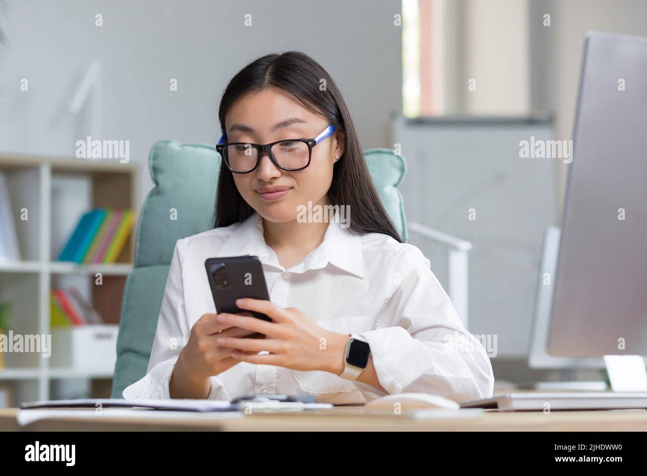 Una giovane bella donna asiatica d'affari in occhiali sta tenendo un telefono nelle sue mani, componendo un messaggio. Seduto su una scrivania in un ufficio moderno, guardando la fotocamera Foto Stock