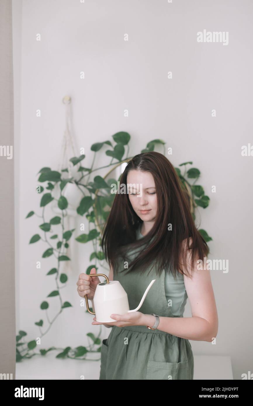 Giovane donna in grembiule, casalinga o giardiniere si prende cura della pianta verde, innaffiare vasi di fiori in camera accogliente. Concetto di giardinaggio e hobby Foto Stock