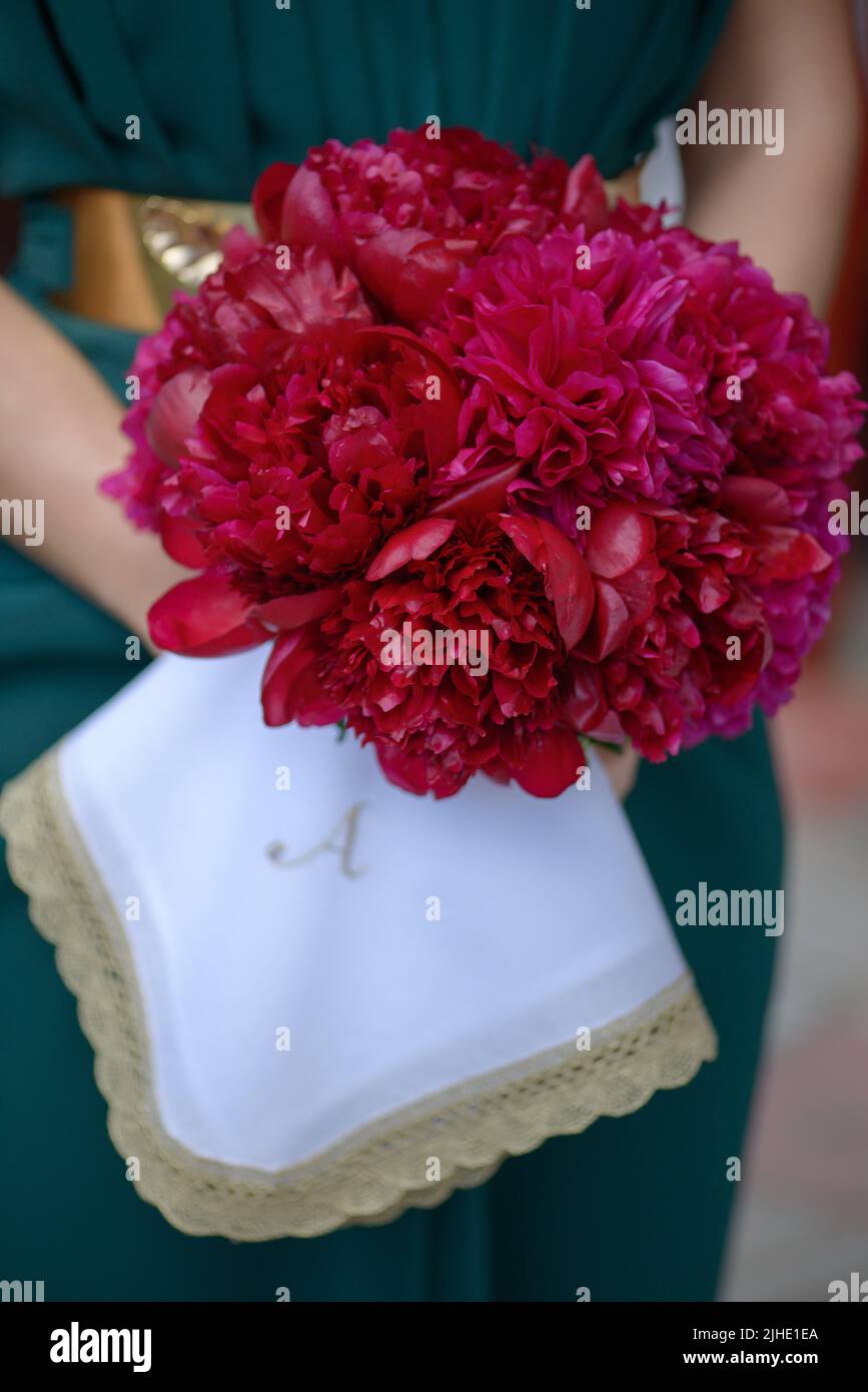 Donna in vestito verde con cintura dorata di grandi dimensioni con bouquet di peonie rosse fringy e un panno ricamato monogramato con lettera A. Foto Stock