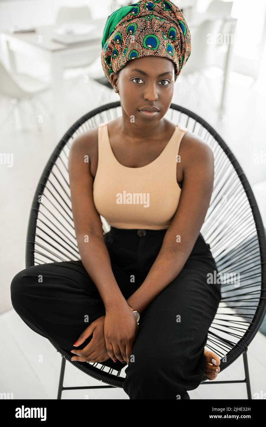 Ritratto di giovane donna alla moda Black African. Seduta su una sedia moderna guardando seriamente in macchina fotografica indossando la tradizionale sciarpa Africana testa Foto Stock