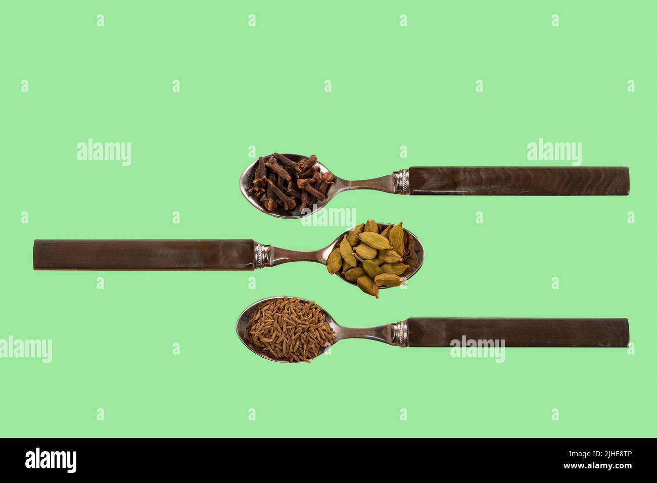 selezione di spezie indiane assortite semi di cumino cardamomo cardamon cialde chiodi di garofano su cucchiai su un colorato sfondo verde Foto Stock