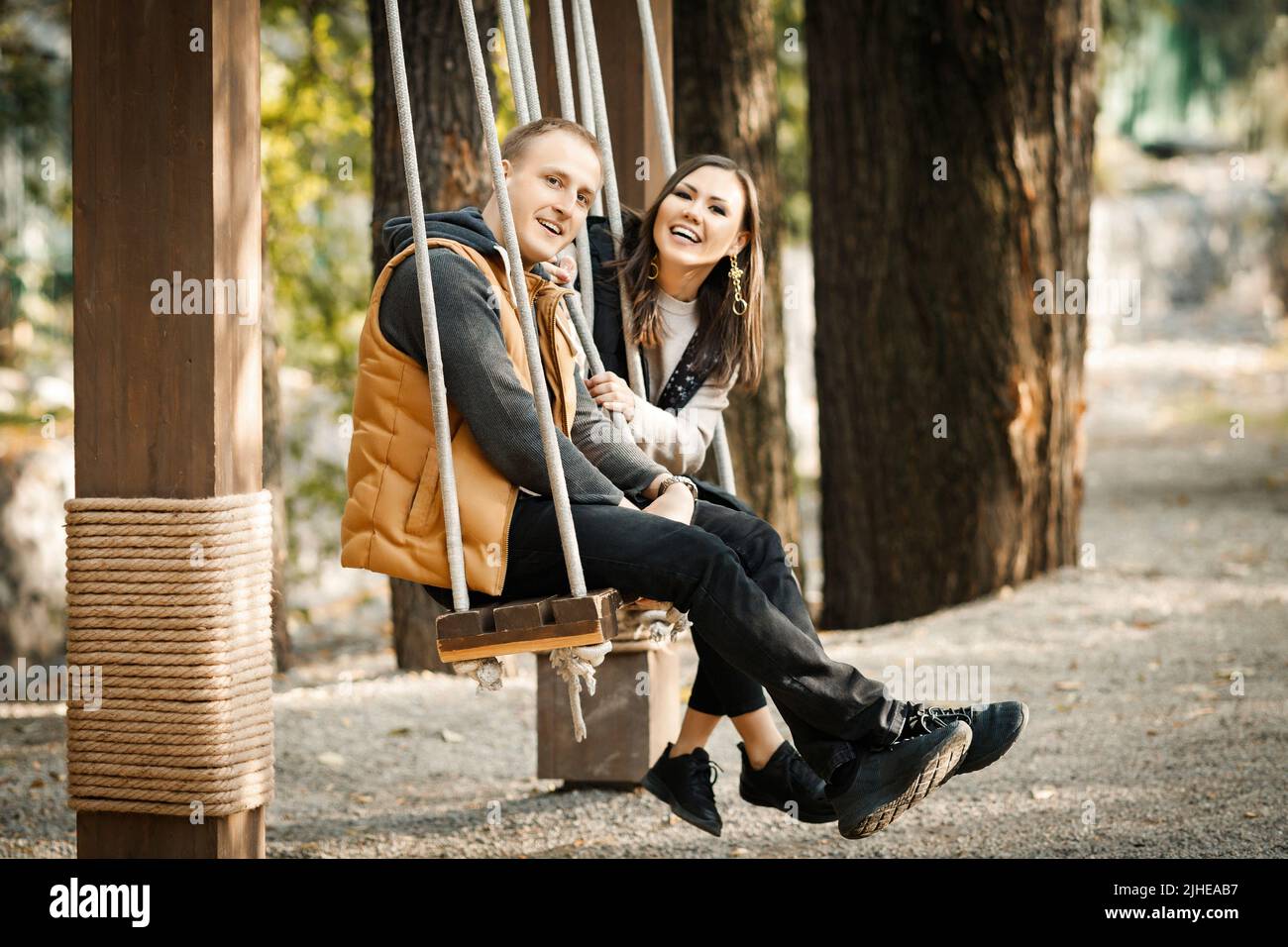Vista laterale di giovane coppia carino in sella a swing in autunno in abiti caldi di colori di base nel parco all'aperto Foto Stock