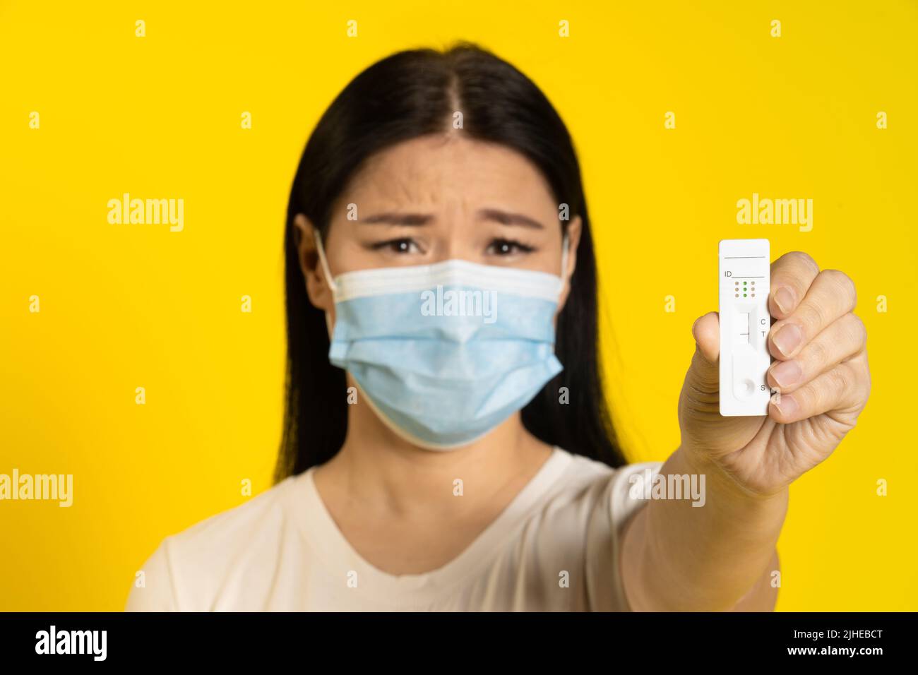 Donna asiatica in maschera medica e test in mano triste guardando la fotocamera isolato su sfondo giallo. Concetto pandemico, varicella o coronavirus scimmia, stile di vita sano. Attenzione selettiva al test. Foto Stock