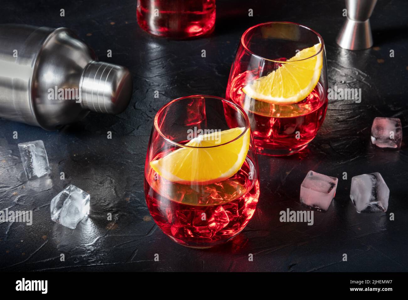 Aperitivo Campari guarnito con arance fresche, con un cocktail shaker, cubetti di ghiaccio, un bicchiere e una bottiglia sullo sfondo Foto Stock