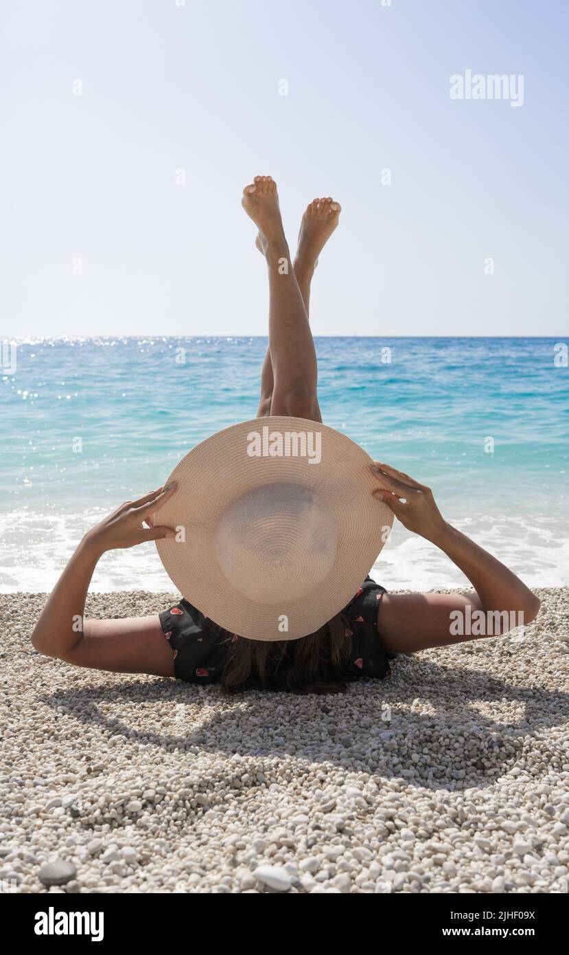 Donna attraente con un cappello di paglia estate sta riposando sulla spiaggia di ciottoli e guardando il mare turchese bello con le gambe sollevate Foto Stock