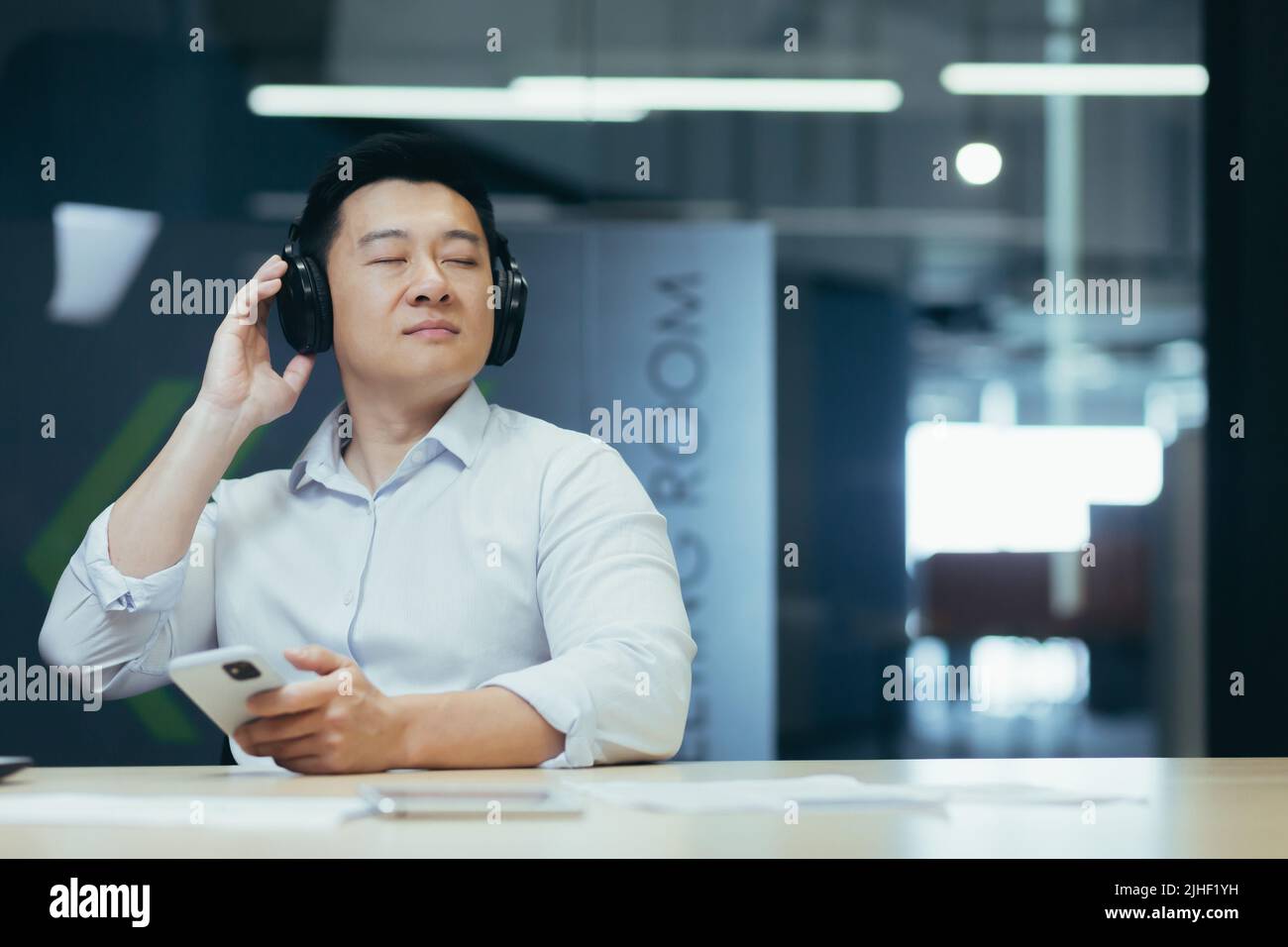 Proprietario di affari del boss asiatico che si rilassa in ufficio, ascoltando musica rilassante e rilassante, uomo seduto alla scrivania, usando le grandi cuffie e l'app di ascolto online sul telefono Foto Stock
