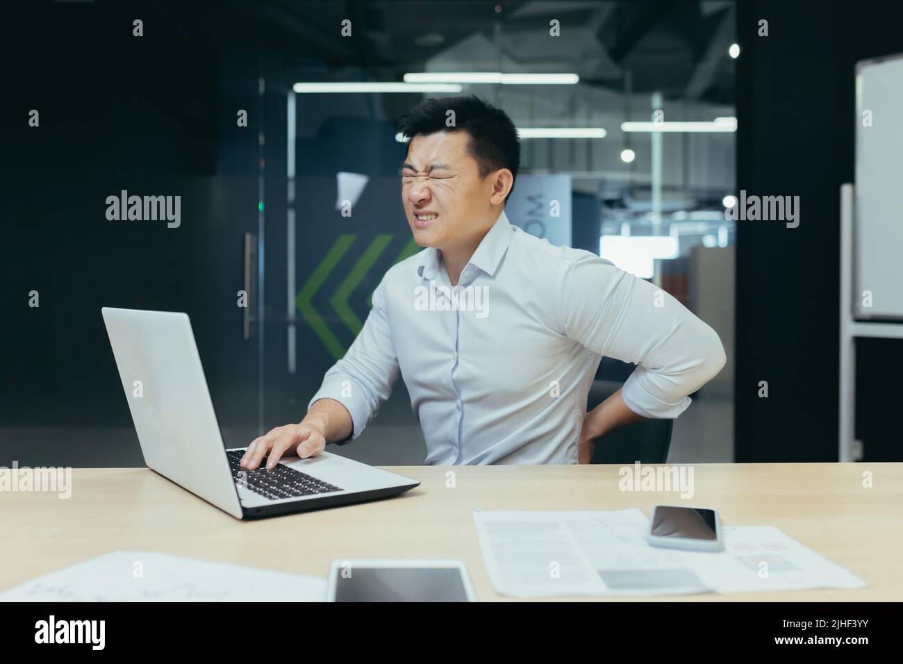 Uomo d'affari asiatico che lavora in ufficio, con grave mal di schiena, lavoratore stanco che lavora con il laptop. Foto Stock