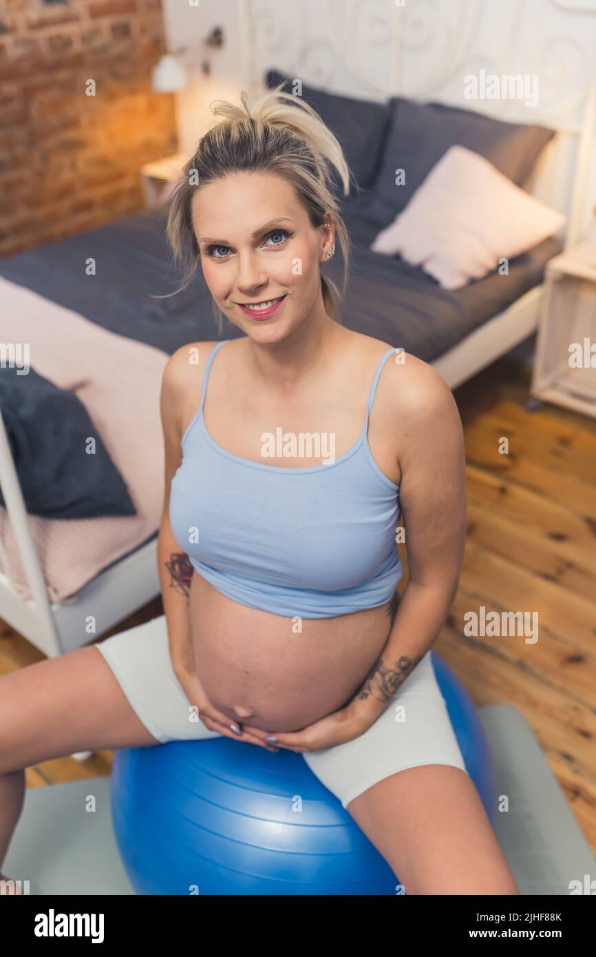 Colpo verticale di una ragazza incinta millenaria caucasica in una cima di serbatoio che tocca il suo ventre grande mentre si siede su una sfera di esercitazione. Foto di alta qualità Foto Stock