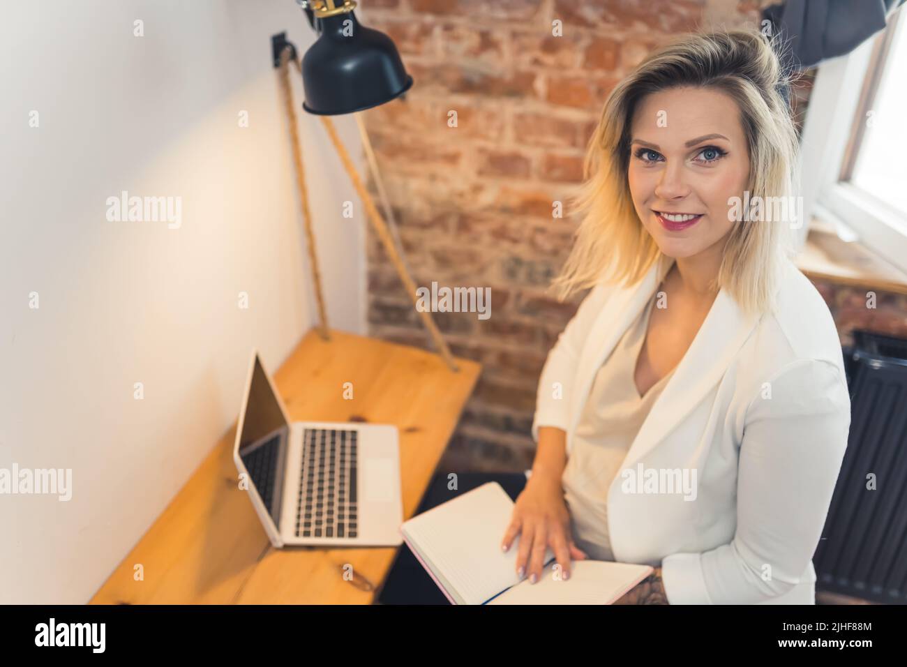 Donna d'affari caucasica professionista incinta in un blazer bianco che guarda la macchina fotografica mentre si siede alla sua scrivania d'ufficio con un computer portatile e un calendario. Foto di alta qualità Foto Stock