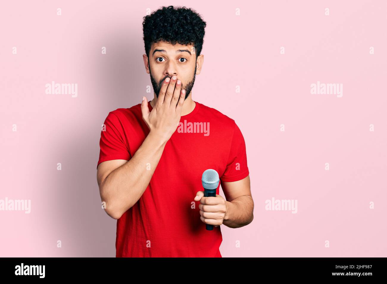 Giovane arabo con la barba che canta il canto usando il microfono che copre la bocca con la mano, scioccato e impaurito per errore. Espressione sorpresa Foto Stock