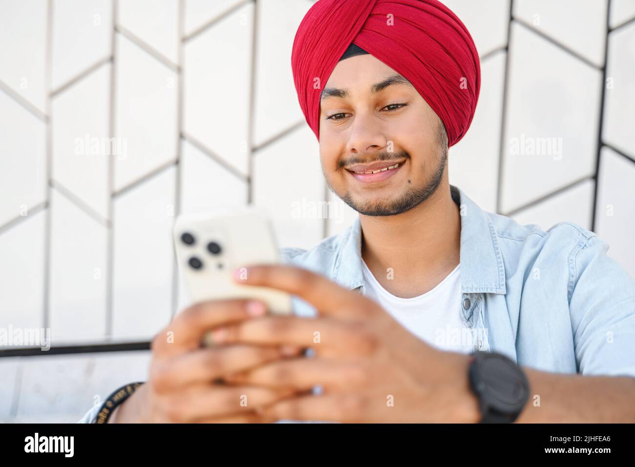 Allegro ragazzo indiano che indossa i tradizionali messaggi turbani, SMS sullo smartphone. Lo studente indù maschile si siede utilizzando l'app mobile all'aperto, scorrimento feed news, websurfing, gioco Foto Stock