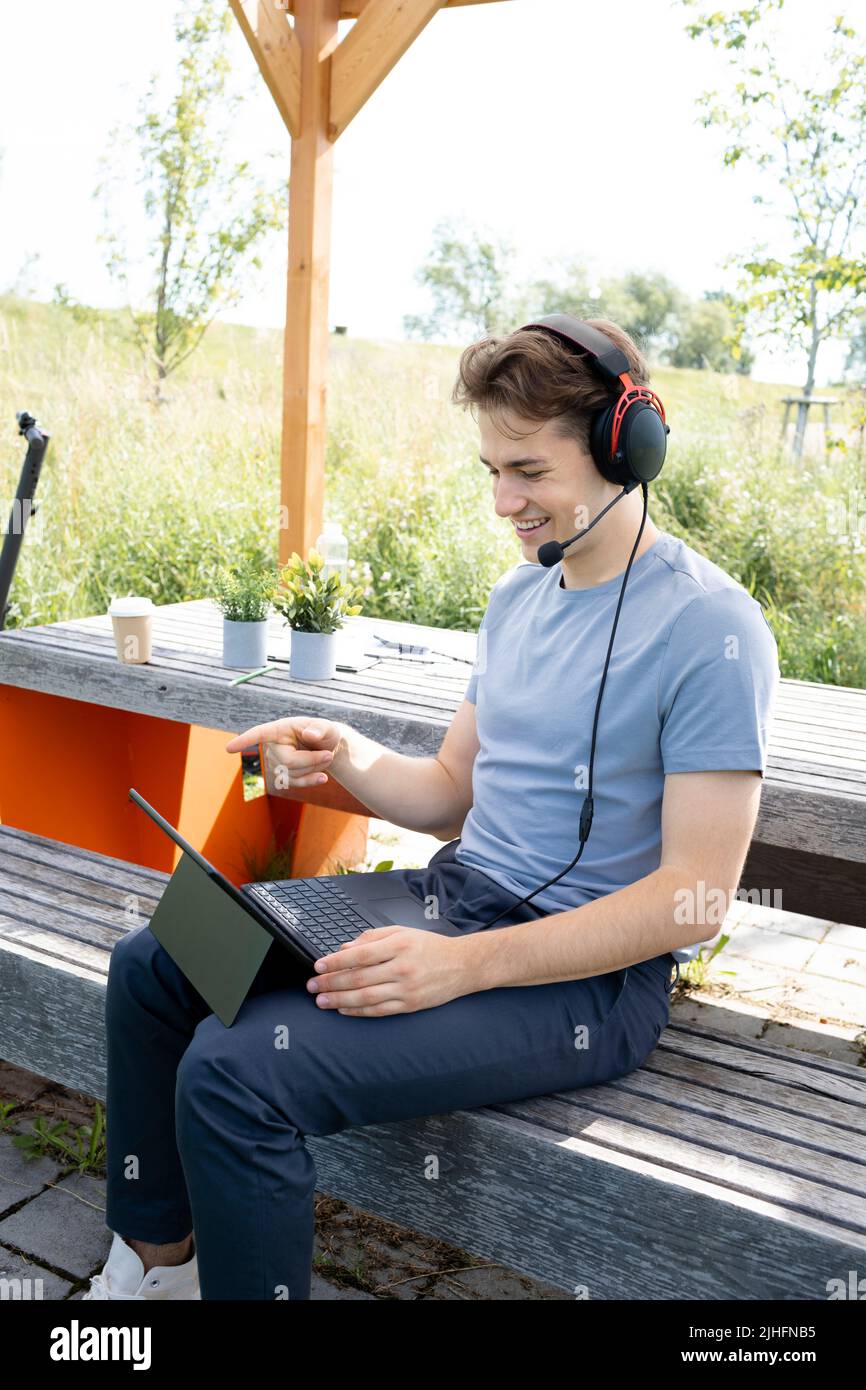giovane e fresco con camicia grigia seduta all'aperto in un parco con cuffie e lavoro sul suo tablet, sullo sfondo e-scooter è in piedi Foto Stock