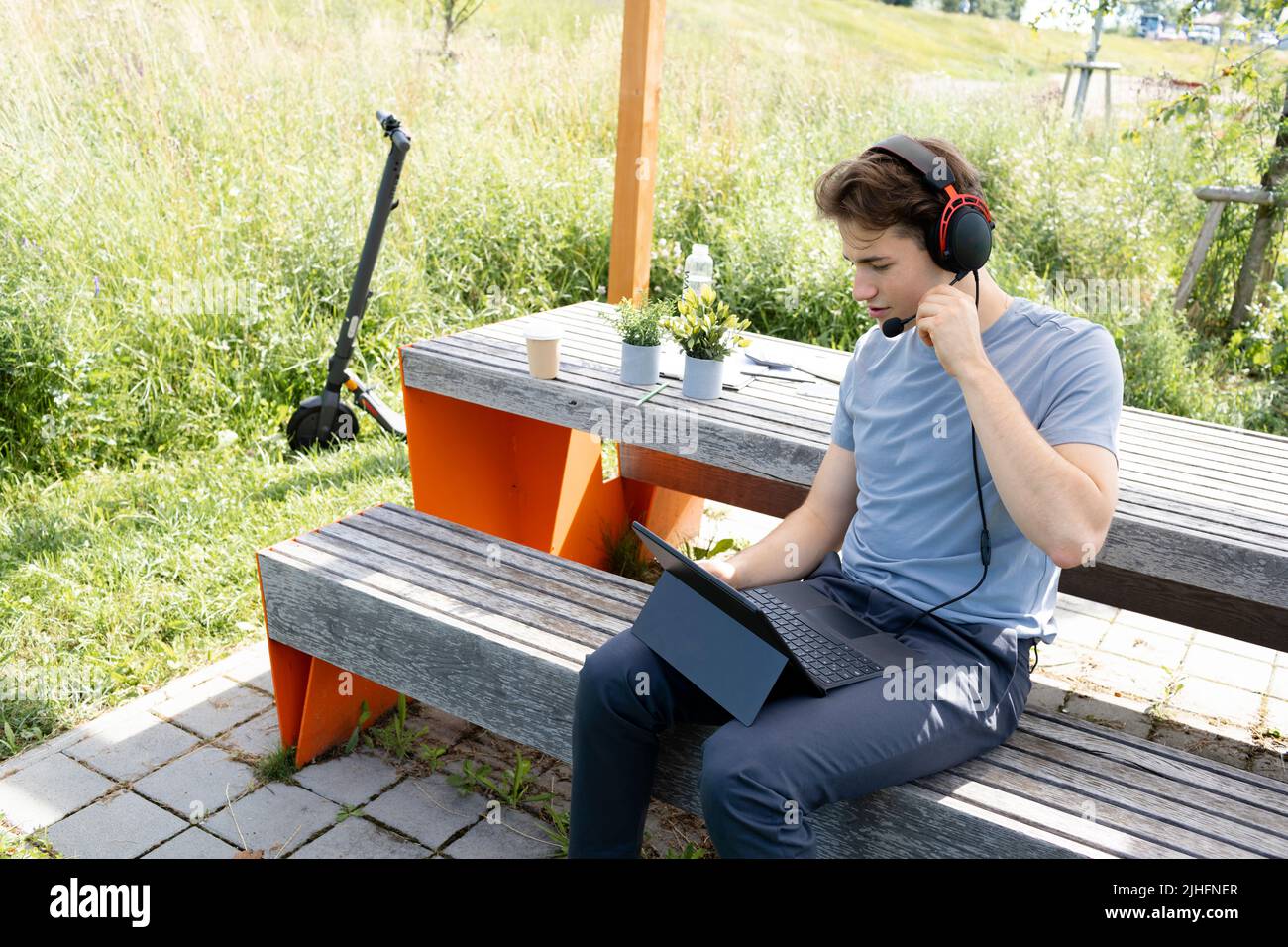 giovane e fresco con camicia grigia seduta all'aperto in un parco con cuffie e lavoro sul suo tablet, sullo sfondo e-scooter è in piedi Foto Stock