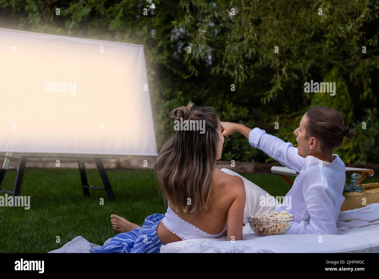 Giovane coppia eterosessuale sititng sul pavimento di erba che punta lo schermo del proiettore mentre nel cinema all'aperto Foto Stock
