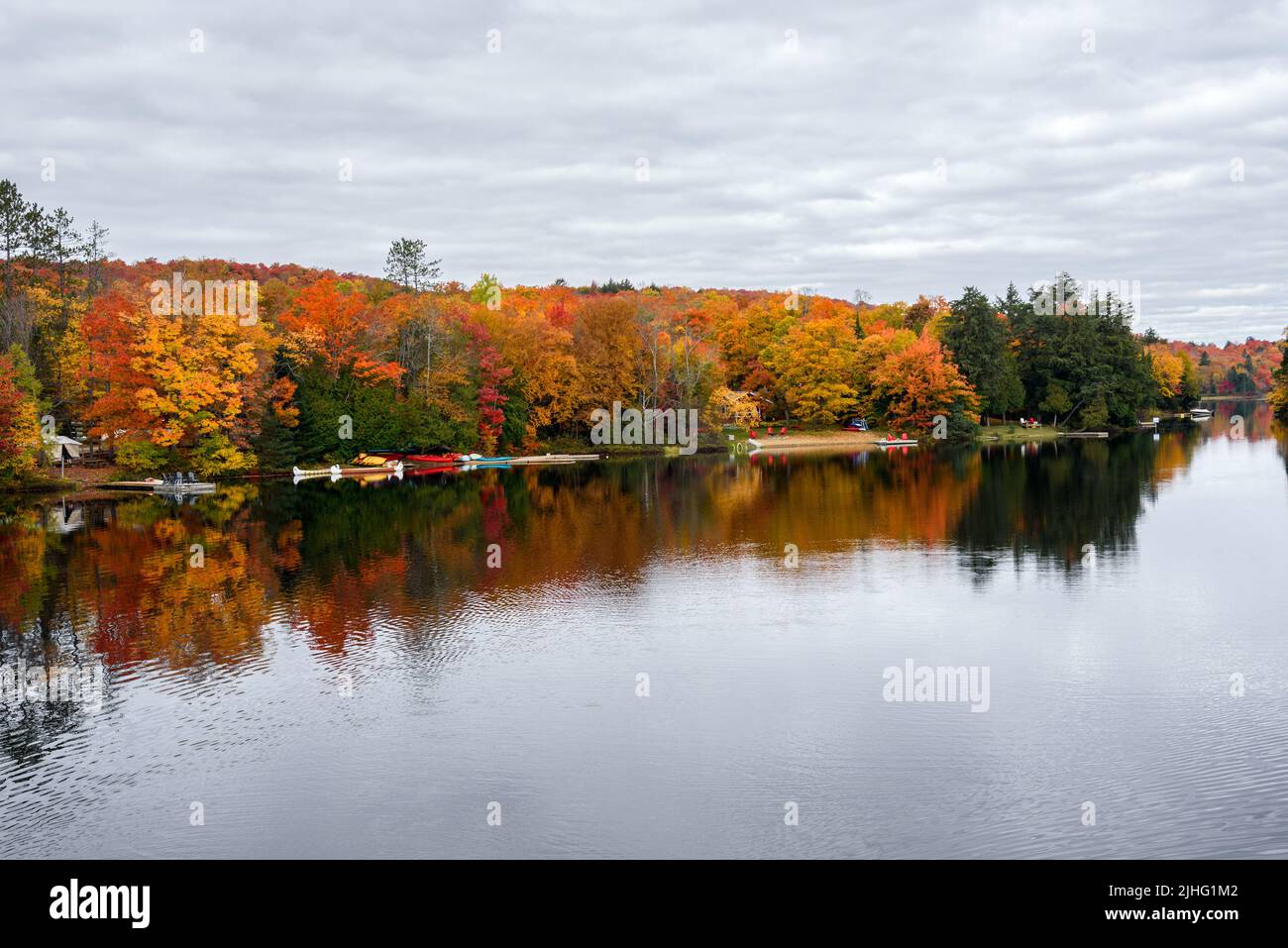 Jetties in legno con sedia adirondack su di loro e canoe colorate lungo la riva boschiva di un lago in una nuvolosa giornata autunnale, incredibile fogliame autunno. Foto Stock