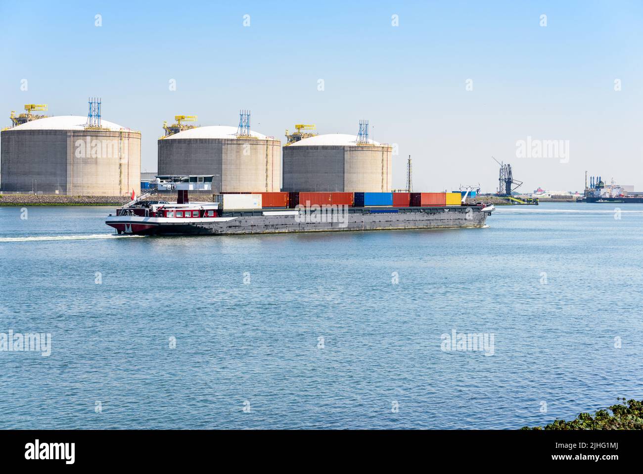 Chiatta contenitore che naviga oltre i serbatoi di GNL in un porto in una giornata estiva limpida Foto Stock