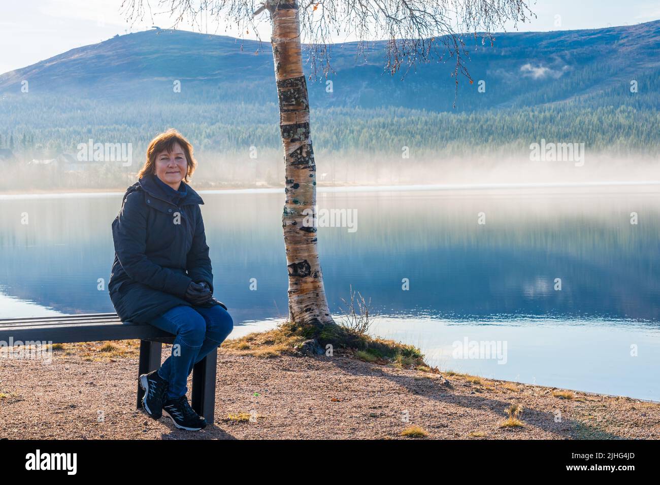 Donna seduta su una panchina in una spiaggia guardando la bella vista con montagna e fiume, Gällivare, Lapponia svedese, Svezia Foto Stock