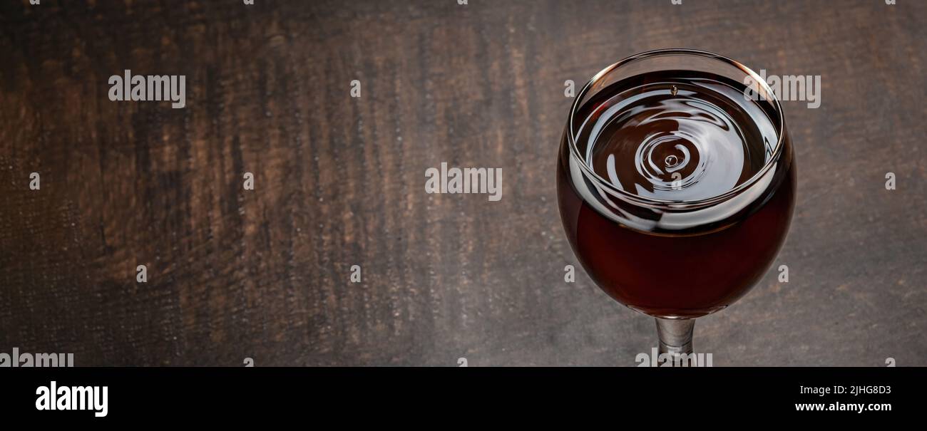 Gocce di vino e onde che si formano in un bicchiere pieno di vino rosso in piedi su un tavolo di legno Foto Stock