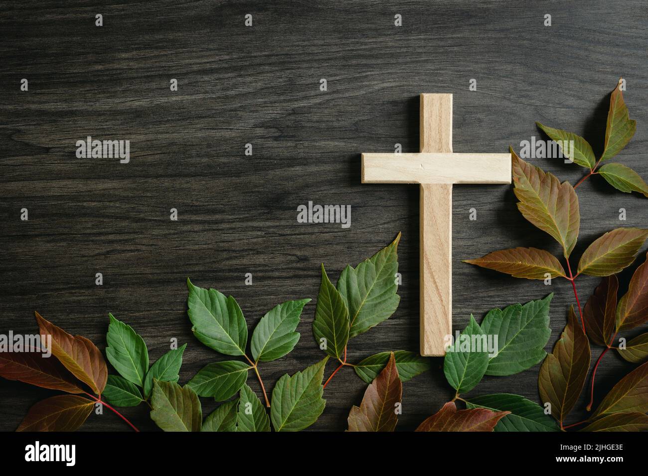 Piccola croce cristiana su sfondo di legno scuro con un bordo di foglie autunnali mutevoli in rosso e verde con spazio copia Foto Stock