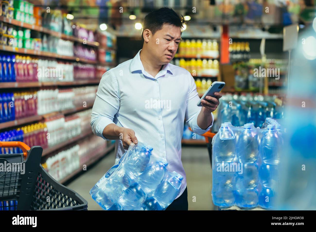 Crisi alimentare, carenza di acqua potabile, uomo asiatico in supermercato acquistare acqua in bottiglia, nervoso e triste lettura cattive notizie al telefono Foto Stock