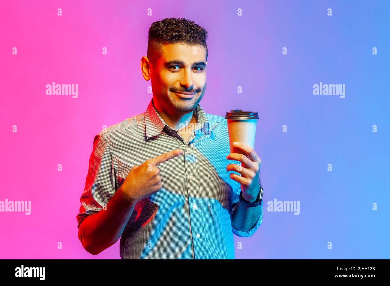 Ritratto di felice uomo ottimista in camicia tenendo il dito puntato verso la tazza di caffè in mano guardando la fotocamera, ha bisogno di energia. Studio interno girato isolato su colorato sfondo di luce al neon. Foto Stock