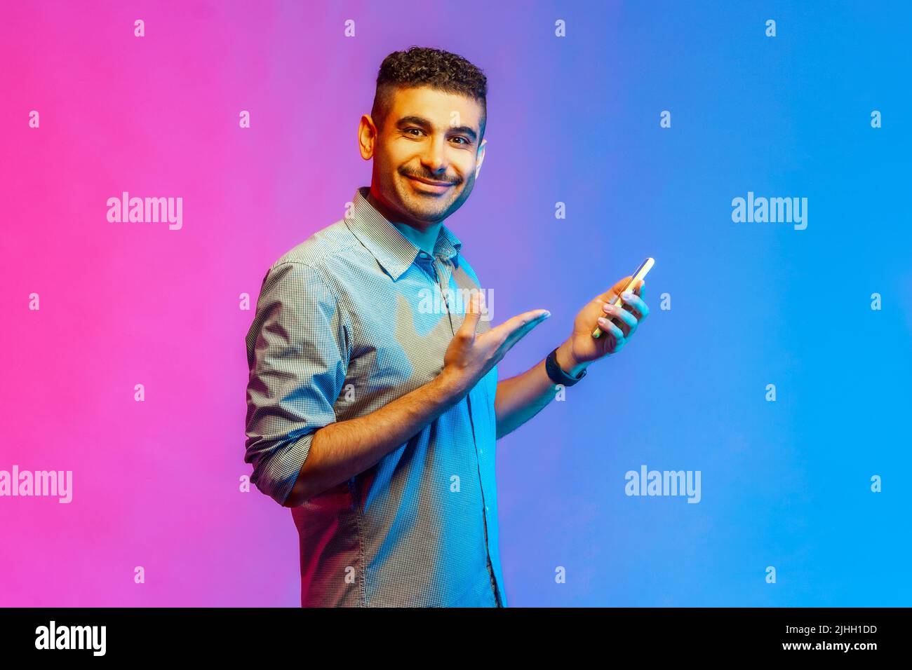 Vista laterale di un uomo ottimista felice in camicia utilizzando il telefono cellulare, sorridendo, mostrando il suo telefono cellulare, godendo l'app mobile. Studio interno girato isolato su colorato sfondo di luce al neon. Foto Stock