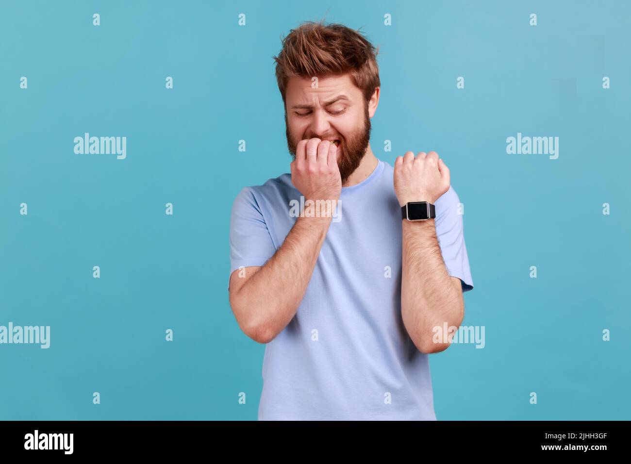 Ritratto di impaziente preoccupato uomo barbuto guardare orologio da polso, avendo ansioso infelice espressione, essere nervoso, troppo tardi, mordere le unghie. Studio interno girato isolato su sfondo blu. Foto Stock