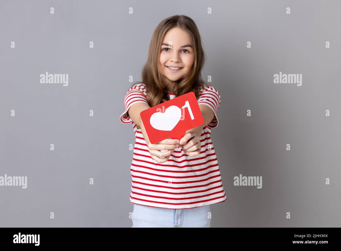 Ritratto di una bambina sorridente che indossa una T-shirt a righe che mostra un'icona a forma di cuore dei social media e guarda la fotocamera con un sorriso carino. Studio interno girato isolato su sfondo grigio. Foto Stock