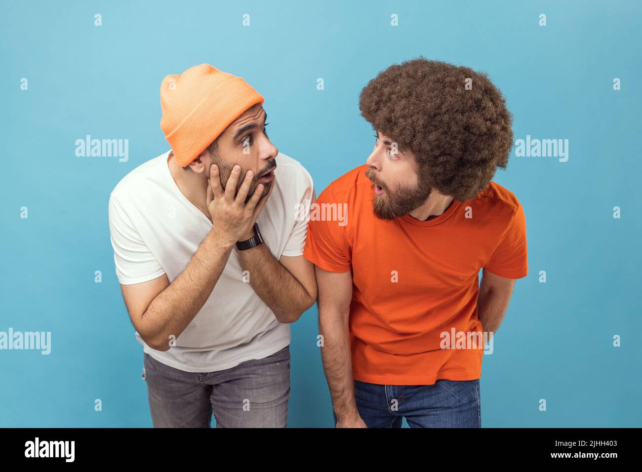 Ritratto di due scioccati stupiti giovani uomini hipster adulti in piedi guardando l'un l'altro con occhi grandi e bocca aperta, essendo sorpreso. Studio interno girato isolato su sfondo blu. Foto Stock